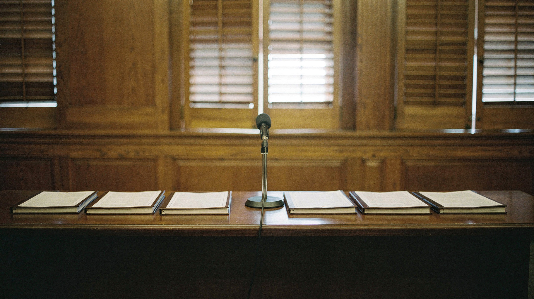 A documentary photograph of a long state capitol press-room table, eight identical bound legal petitions arranged in a neat row, each with a blank cover sheet, a single microphone at the centre, tall shuttered windows behind