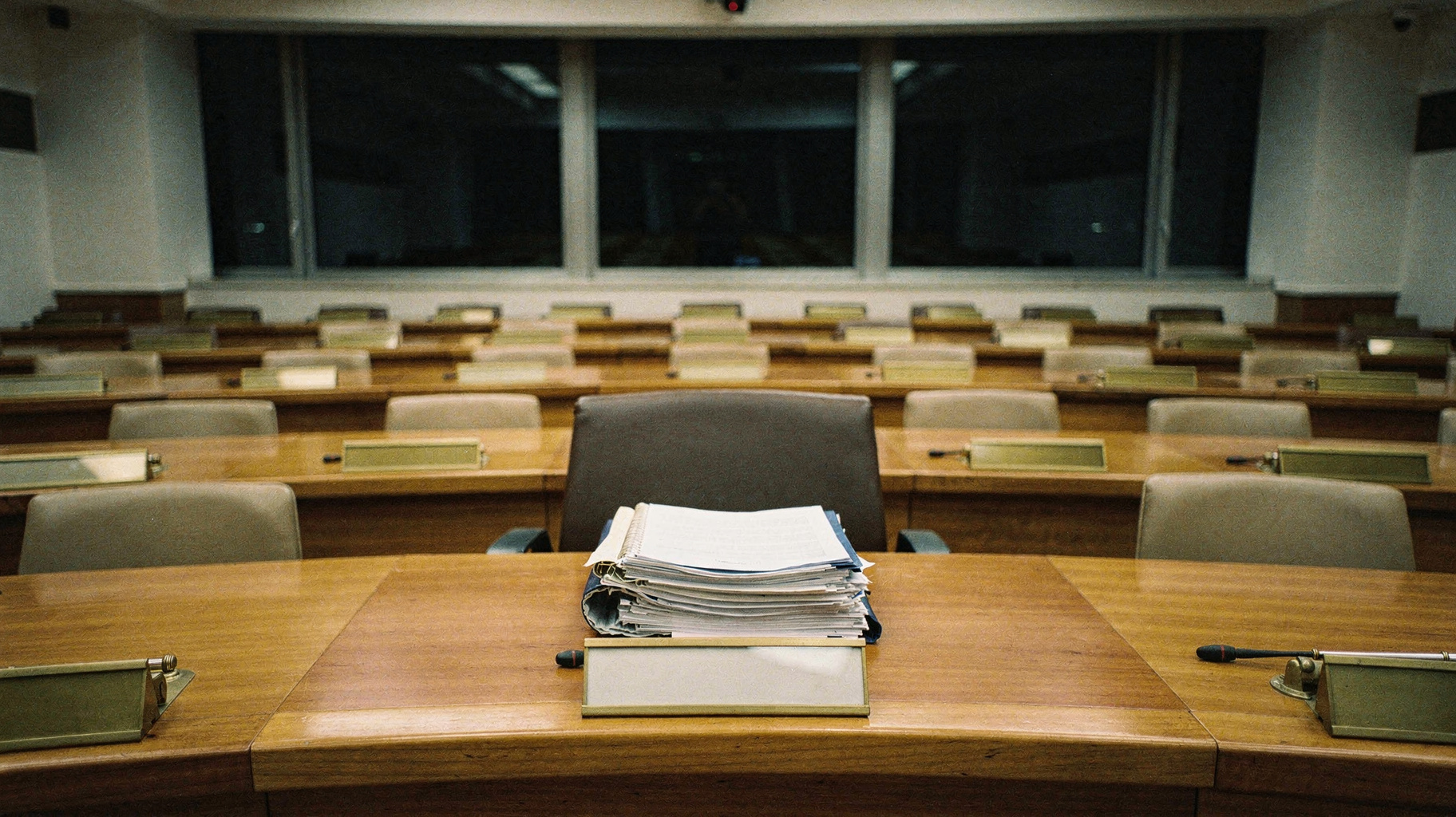 A documentary photograph of an empty European committee room, long curved wooden desks in rows, blank nameplates in brass holders, a thick stack of bound documents left open at the centre seat, tall windows dark behind