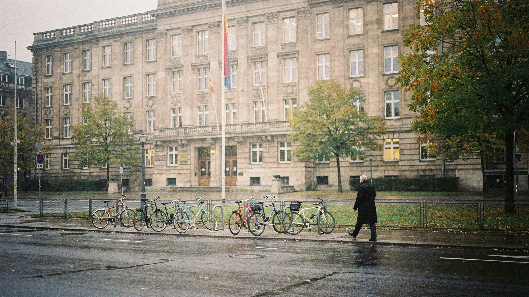 A documentary photograph of a German government ministry building facade on an overcast Berlin morning, a row of bicycles chained at the curb, a single official entering through a side door, muted earth tones