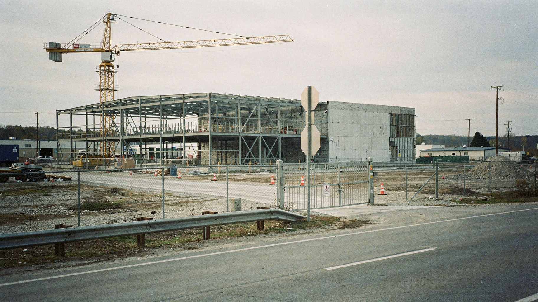 A half-finished data center shell with open steel frame, a crane standing idle beside it, fenced gravel lot in the foreground