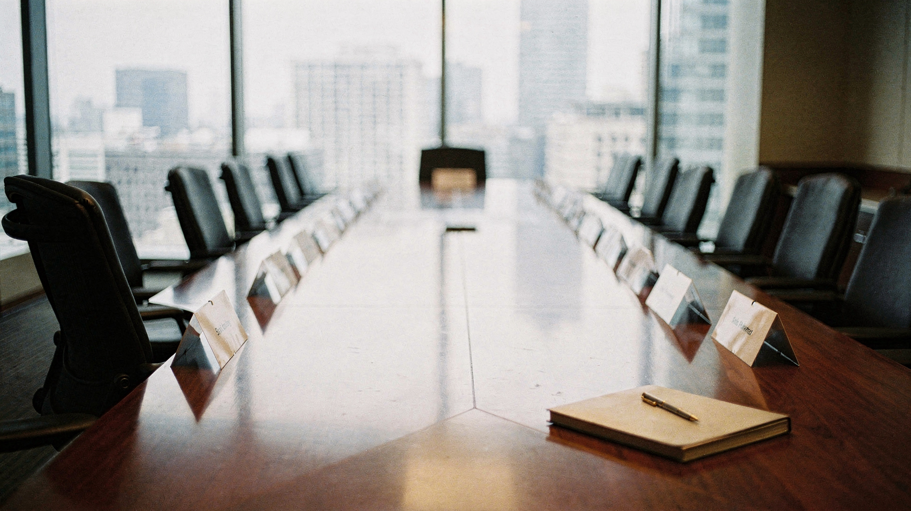A documentary photograph of a formal boardroom with a long polished table, nameplates set out in a row, one chair pulled slightly from the table, city light through tall windows
