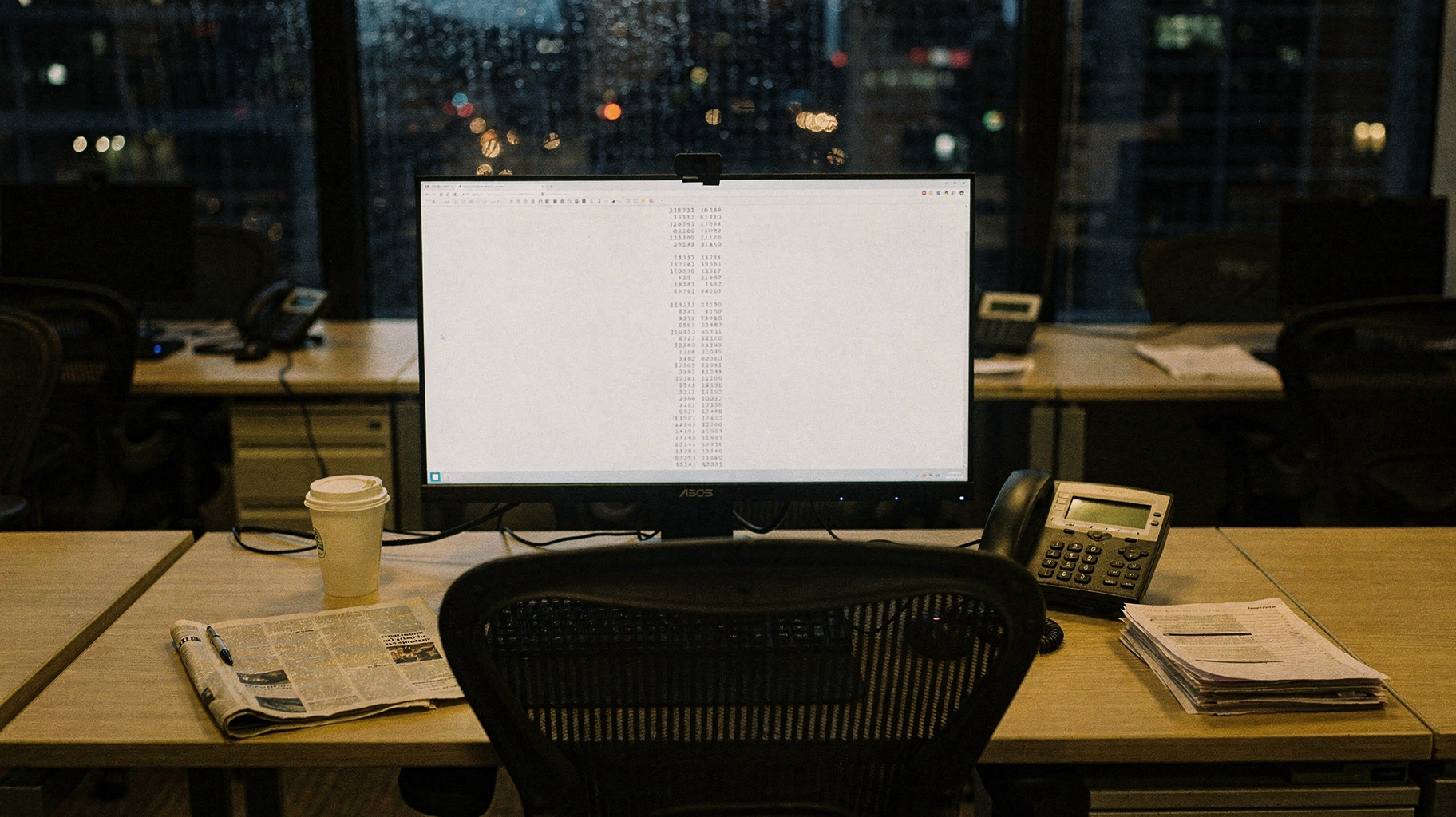 A documentary photograph of a trading desk after hours, a single monitor still lit showing a column of numbers, a paper cup and a folded newspaper, city lights blurred behind rain-streaked glass