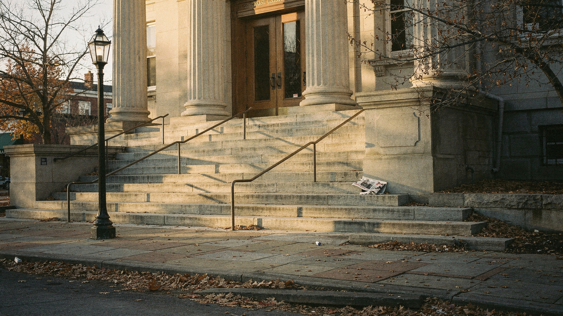 A documentary photograph of a neoclassical courthouse entrance at early morning, fallen leaves gathered on wide stone steps, nobody present, low golden sun raking across the stone