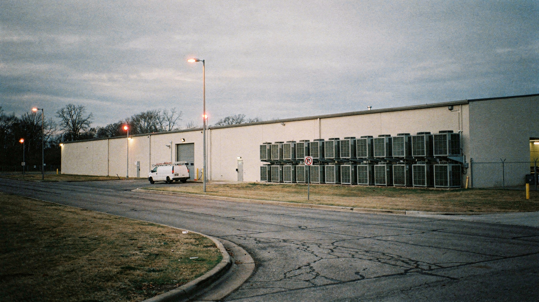 A documentary photograph of a data center exterior at dusk, long low industrial building, cooling units along the wall, a service road, flat grey cloud